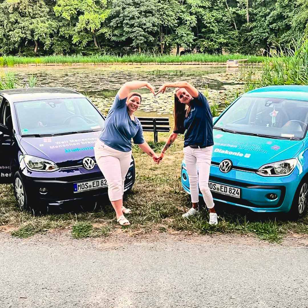Zwei Mitarbeiterinnen stehen vor Ihren Dienstfahrzeugen und bilden mit Ihren Armen ein Herz.